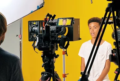 Photograph of young boy being interviewed to camera.  The view is from behind the camera.  The set is yellow and you can see the boys face in the camera's view.
