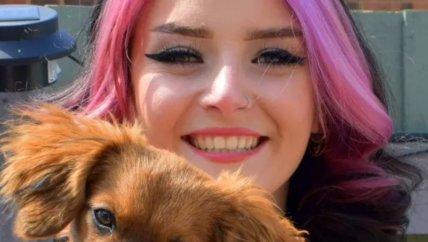 Young woman with pink hair and sun glasses on top of her head smiles at the camera, whilst holding a small dog  