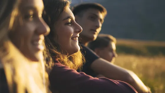 A group of teenagers sit smiling in a row outside in the low light of sunset.