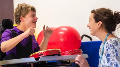 Young boy sat with a red tambourine and across from an adult woman. Both individuals are smiling at each other.