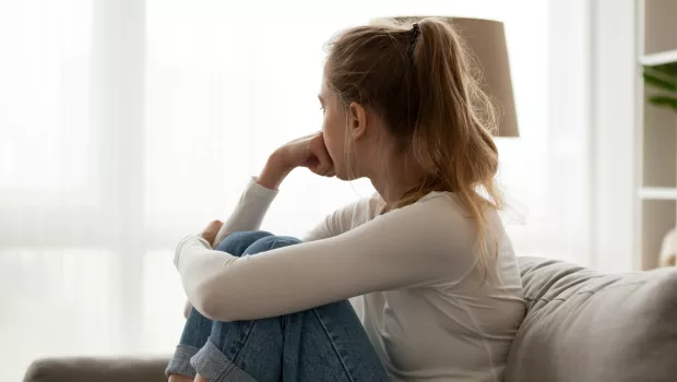 A woman sits on a sofa looking out of the window in a thoughtful, sombre pose.