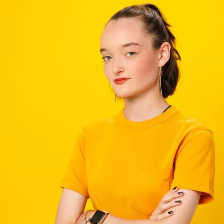 A young woman, Bea, wears yellow against a yellow background with her arms crossed.