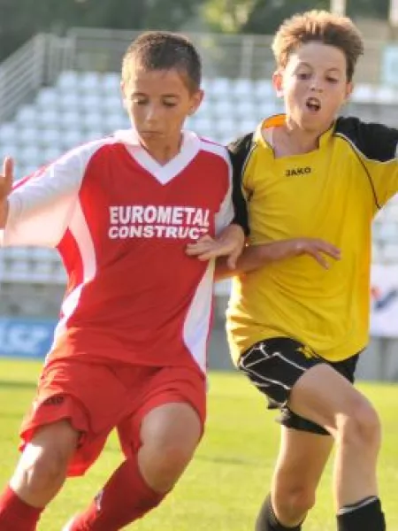 Two young boys wearing football kits running together on a football pitch. They are shoulder-barging each other and running towards the viewer.