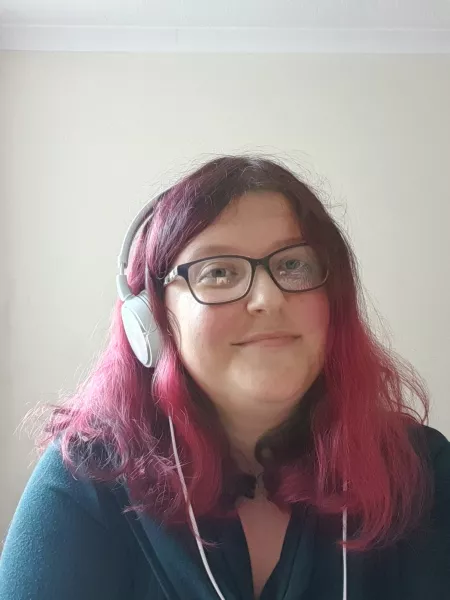 A young woman with red hair smiles at the camera with headphones on.
